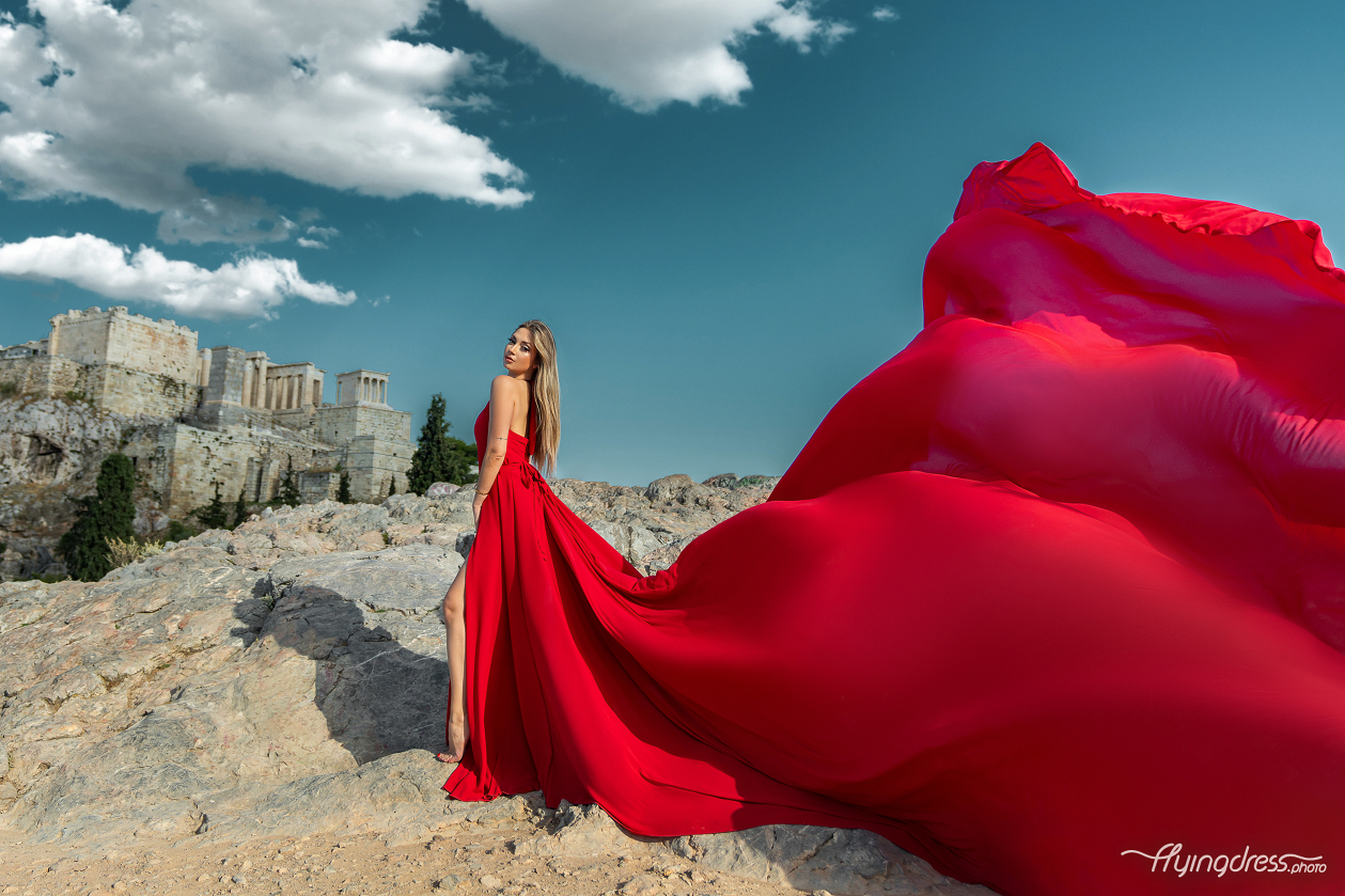 A woman in a flowing red dress poses dramatically on a rocky terrain with the ancient ruins of Athens, including the Acropolis, in the background against a partly cloudy sky.